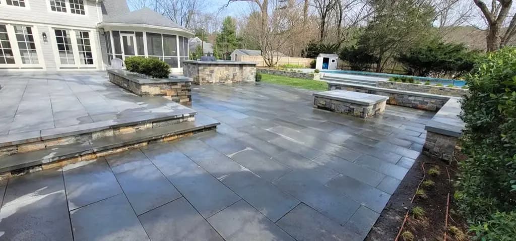 Large gray stone patio with steps, low walls, shrubs, and a swimming pool